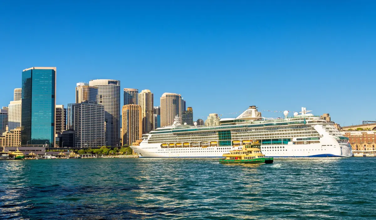 Un grand paquebot blanc amarré devant la skyline de Sydney par une journée ensoleillée, avec un ferry vert et jaune au premier plan