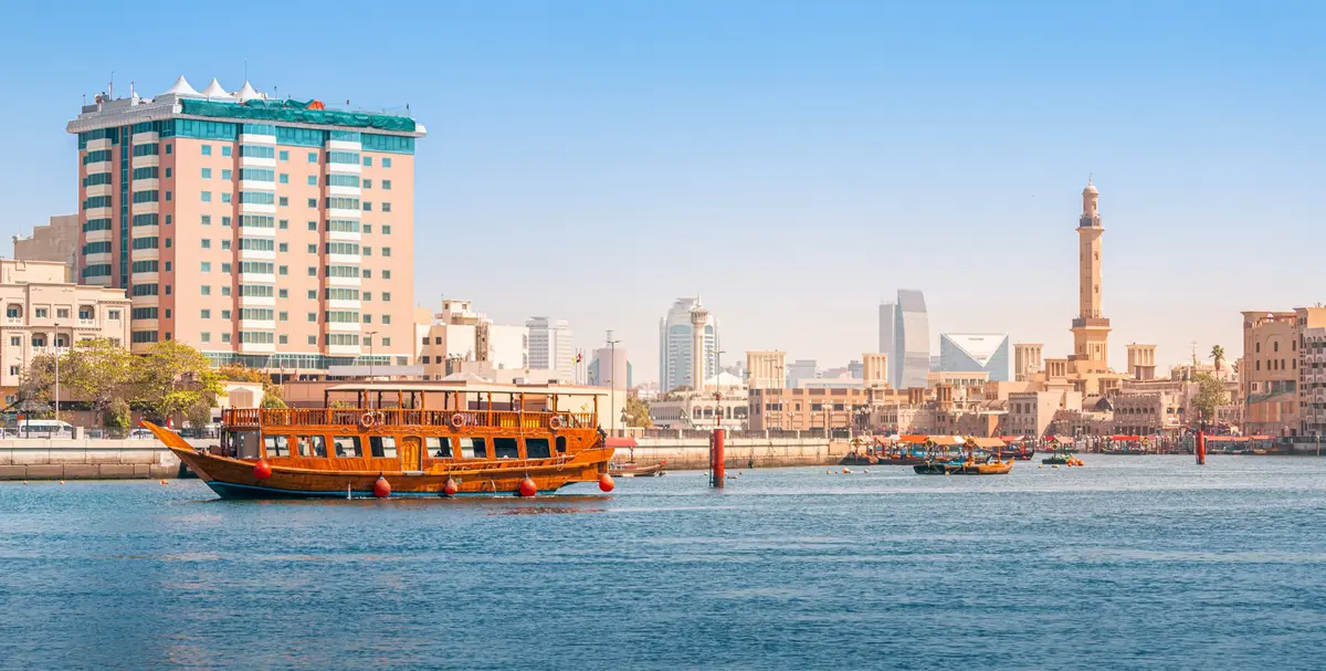 A traditional wooden dhow cruising on Dubai Creek with the old Deira skyline and a minaret behind