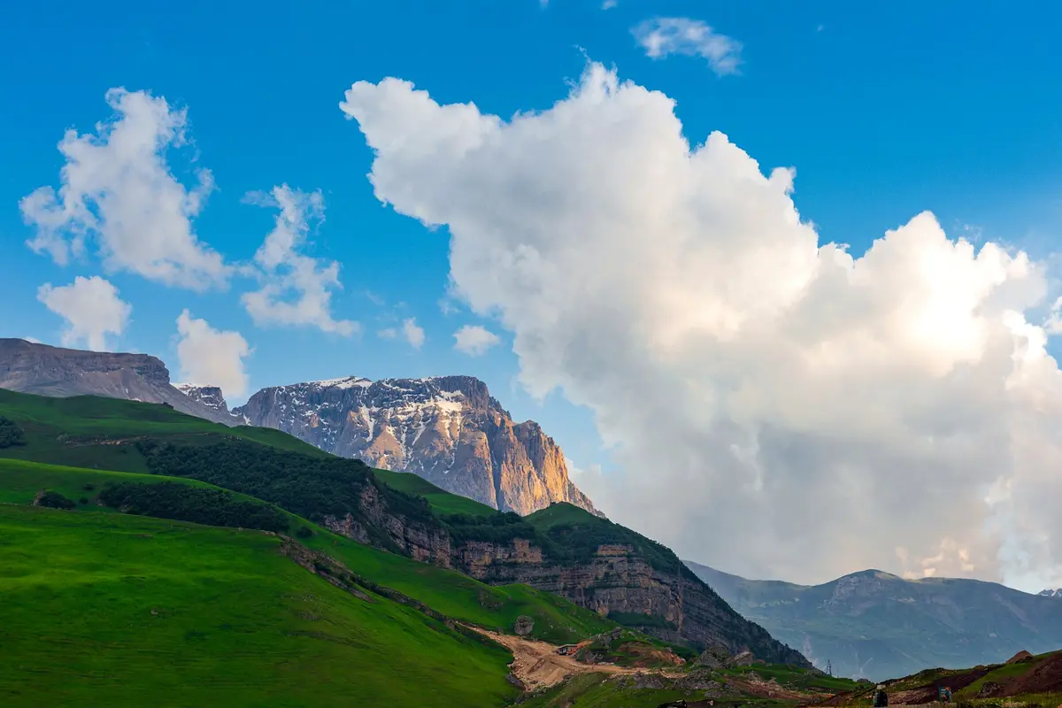 Une montagne rocheuse avec des plaques de neige au-dessus de prairies vertes, sous un ciel bleu profond et de gros nuages