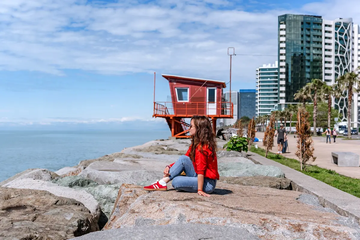 Eine Frau sitzt auf den Felsen am Meer in Batumi, dahinter ein roter Rettungsturm und die Skyline
