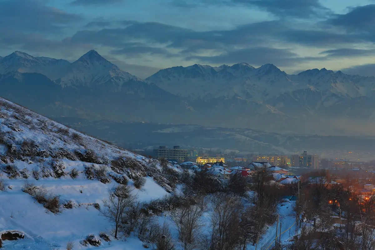 Verschneiter Hang in Almaty in der Dämmerung mit Lichtern der Stadt und den Alatau-Gipfeln dahinter