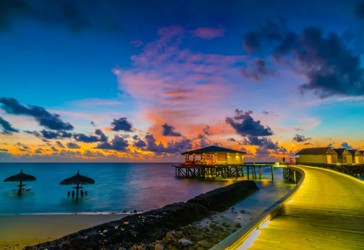 Un ponton en bois courbe et éclairé au coucher du soleil, avec des parasols de paille sur une plage des Maldives