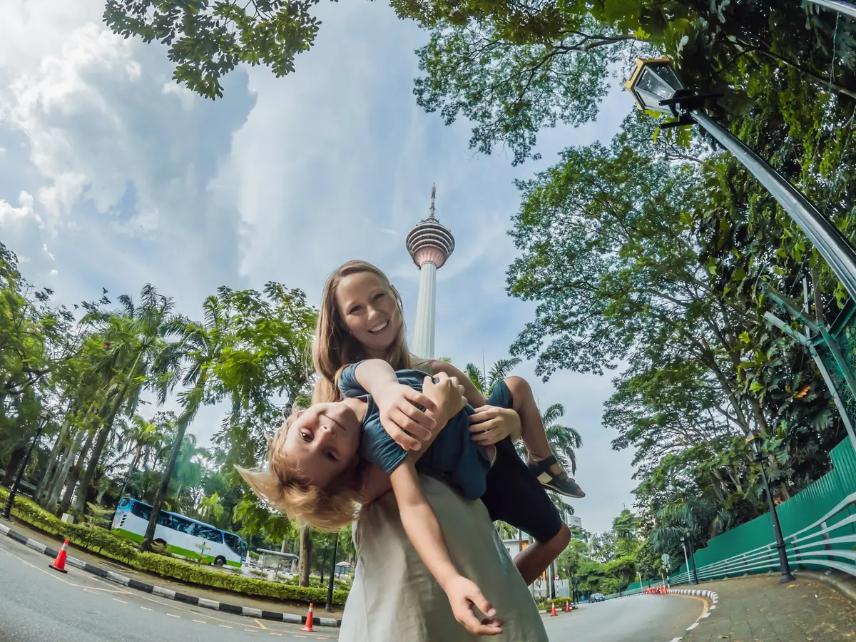 Une mère tient son fils souriant la tête en bas devant la tour de Kuala Lumpur au milieu d'arbres tropicaux