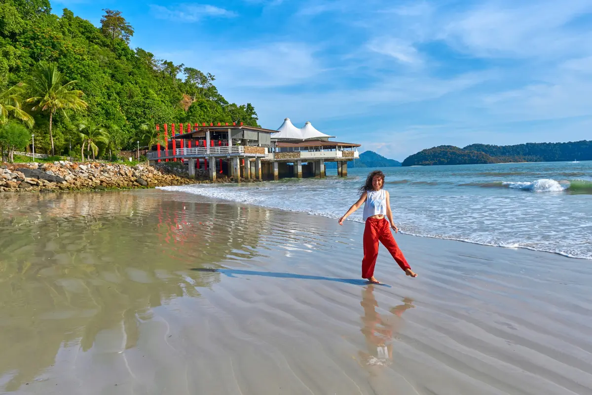 Une femme souriante marche pieds nus sur une plage de sable à Langkawi, avec des îles vertes au loin