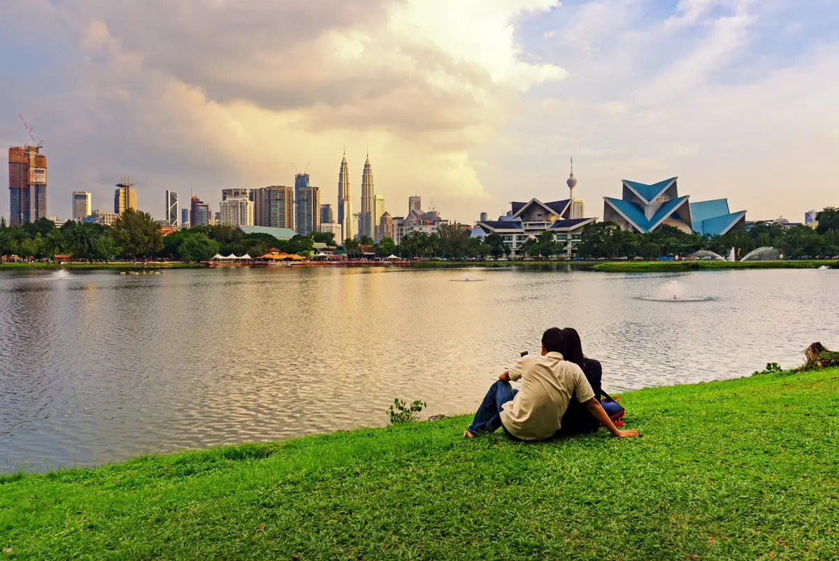 Un couple assis sur la pelouse d'un lac au coucher du soleil face à la silhouette de Kuala Lumpur et aux tours Petronas