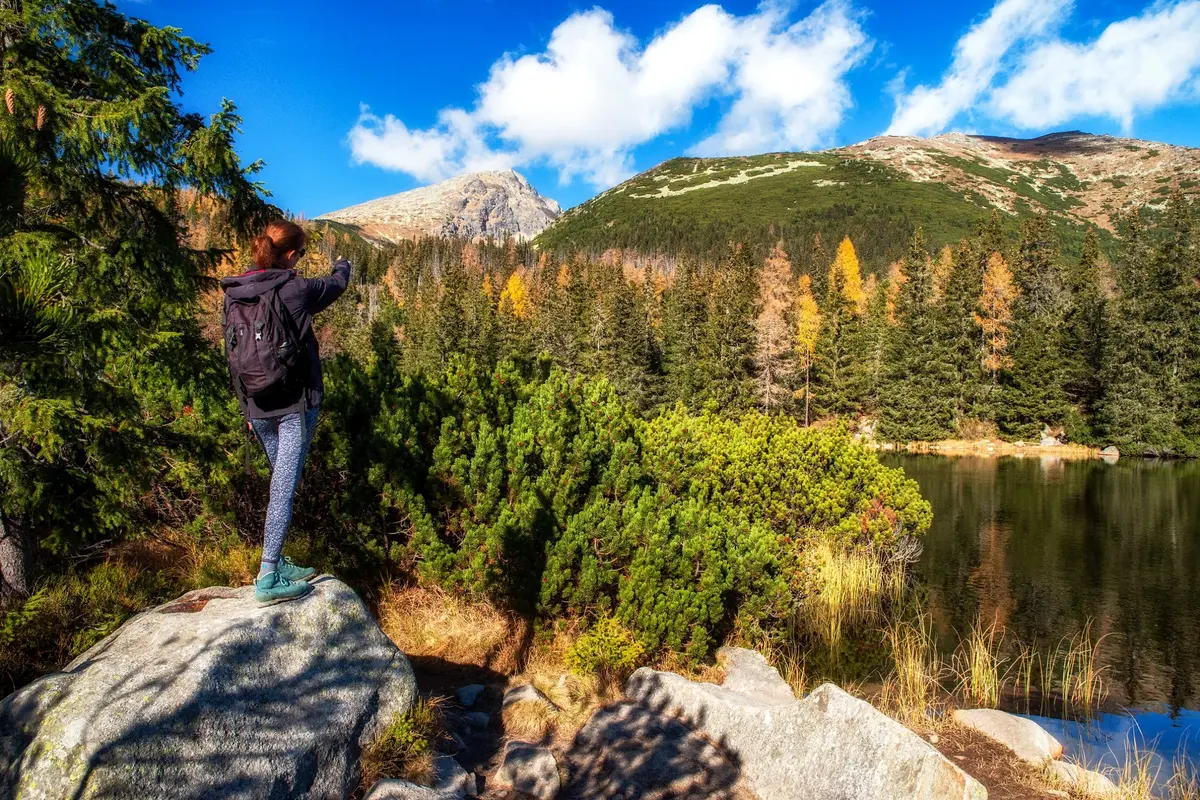 Eine Wanderin zeigt auf einen felsigen Gipfel über einem Bergsee in der Hohen Tatra, zwischen herbstlichen Lärchen