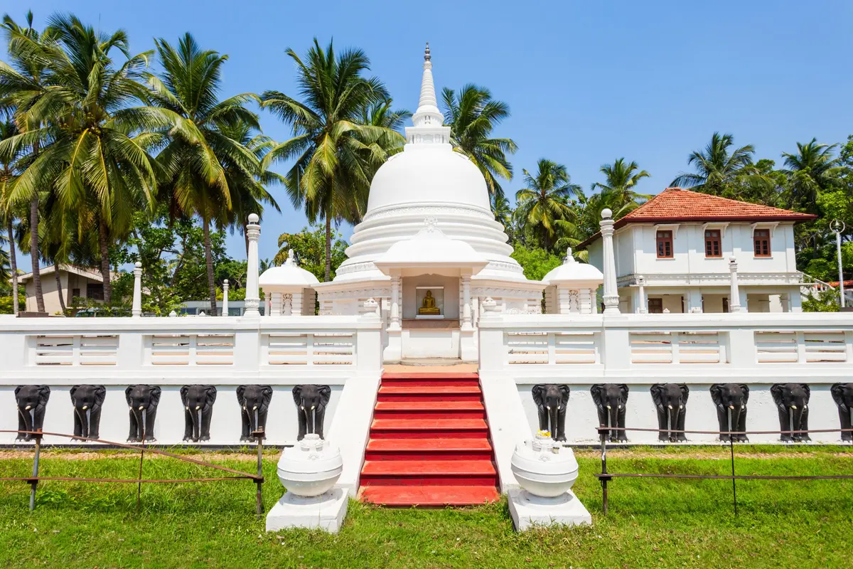 Ein weißer buddhistischer Tempel mit Stupa-Kuppel, roten Stufen und einer mit schwarzen Elefantenfiguren geschmückten Mauer zwischen Palmen in Sri Lanka