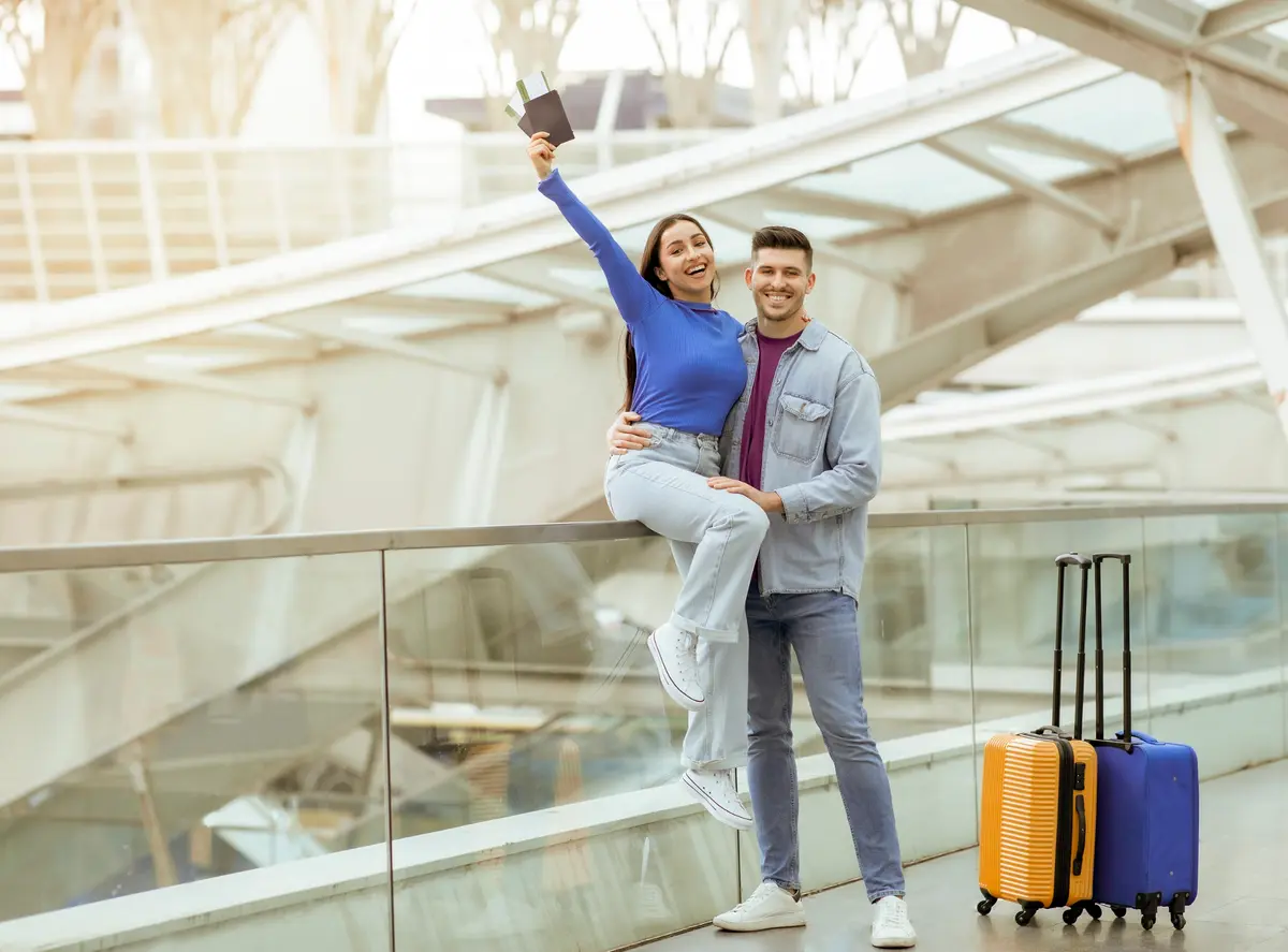 Ein glückliches Paar am Flughafen, die Frau hält Reisepässe und Bordkarten neben zwei Koffern hoch