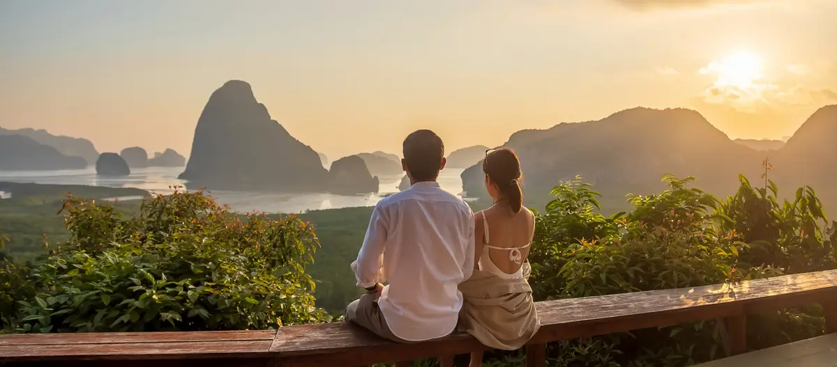 Ein Paar sitzt zusammen und sieht den Sonnenaufgang über Kalksteinfelsen und einer ruhigen Bucht in Phang Nga