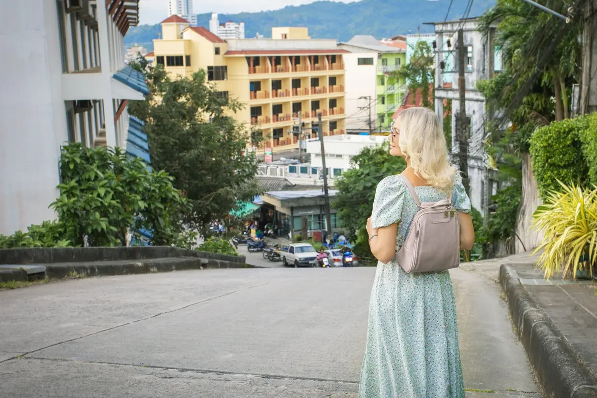 Eine Frau mit Rucksack blickt in Phuket eine abfallende Straße zwischen bunten Gebäuden hinunter