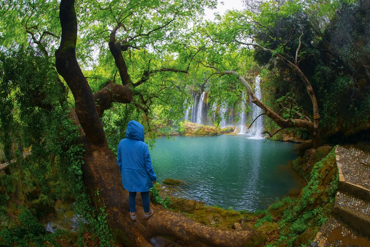 Eine Person im blauen Regenmantel blickt in einem grünen Wald der Region Antalya auf einen Wasserfall und ein türkisfarbenes Becken