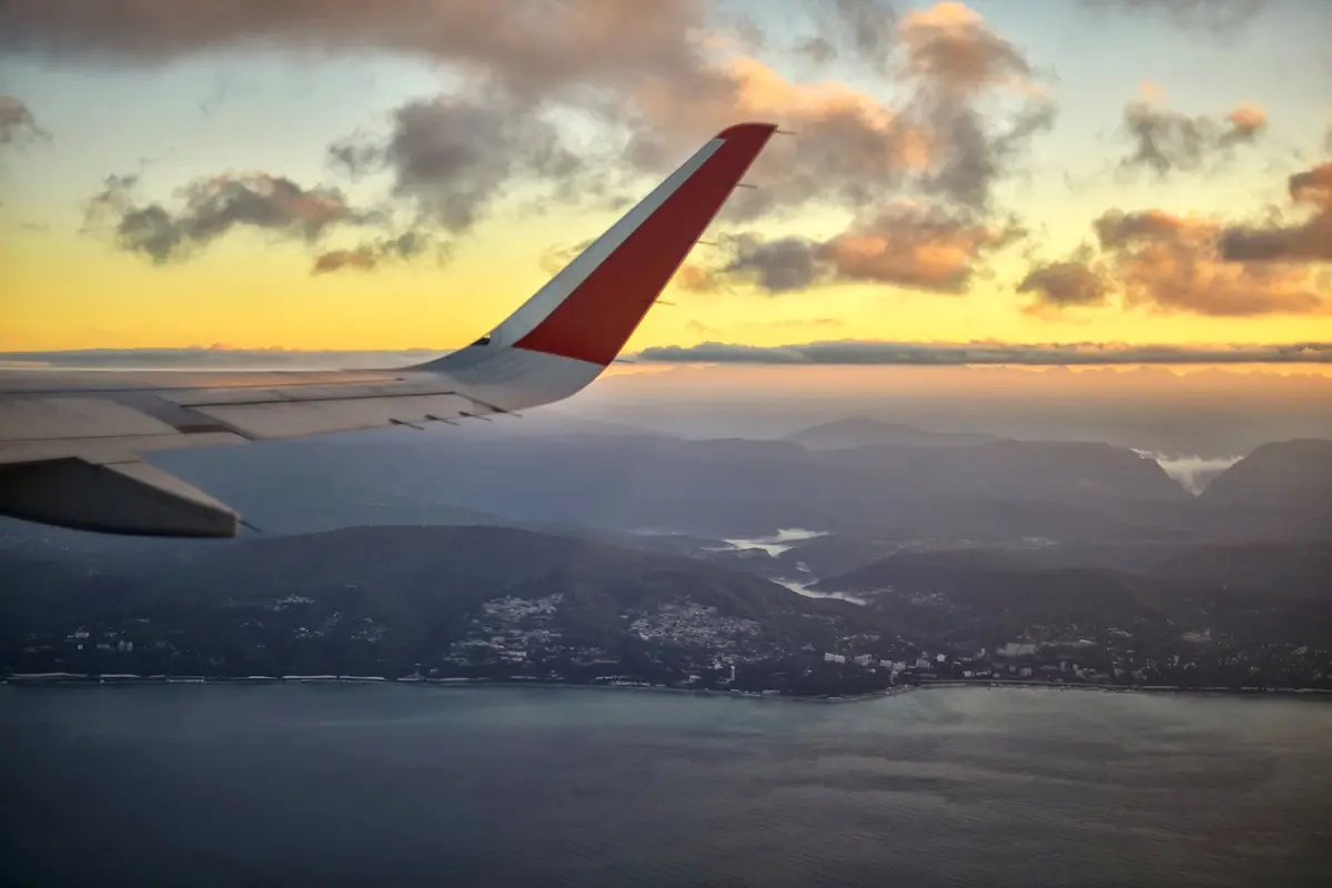 Ein Flugzeugflügel bei Sonnenuntergang über Wolken und einer Küste mit Bergen
