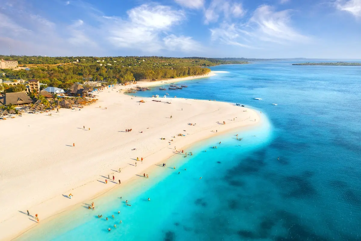 Luftaufnahme eines weißen Sandstrandes auf Sansibar mit Badenden, Fischerbooten und grüner Küste unter blauem Himmel