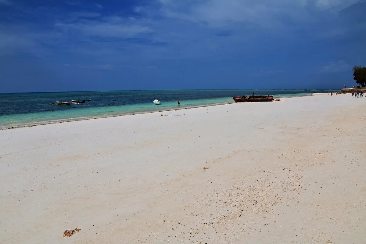 Ein langer weißer Sandstrand an der Küste Sansibars mit türkisfarbenem Flachwasser, Fischerbooten und dunkelblauem offenem Meer