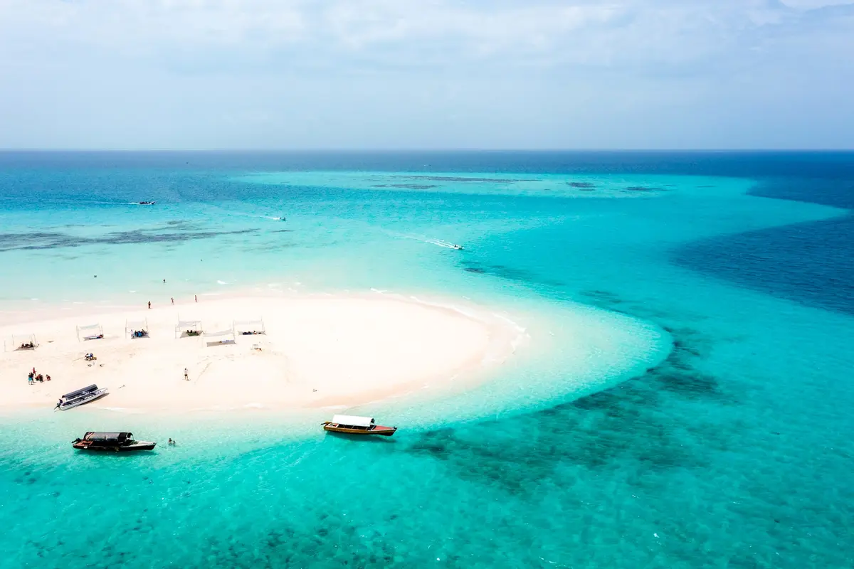 Luftaufnahme einer weißen Sandbank im türkisfarbenen Wasser mit kleinen Booten und Sonnensegeln vor Sansibar